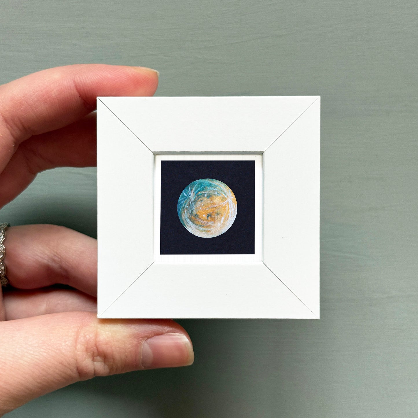 A hand is holding a small white square frame containing a miniature representation of the Earth, with a dark background.