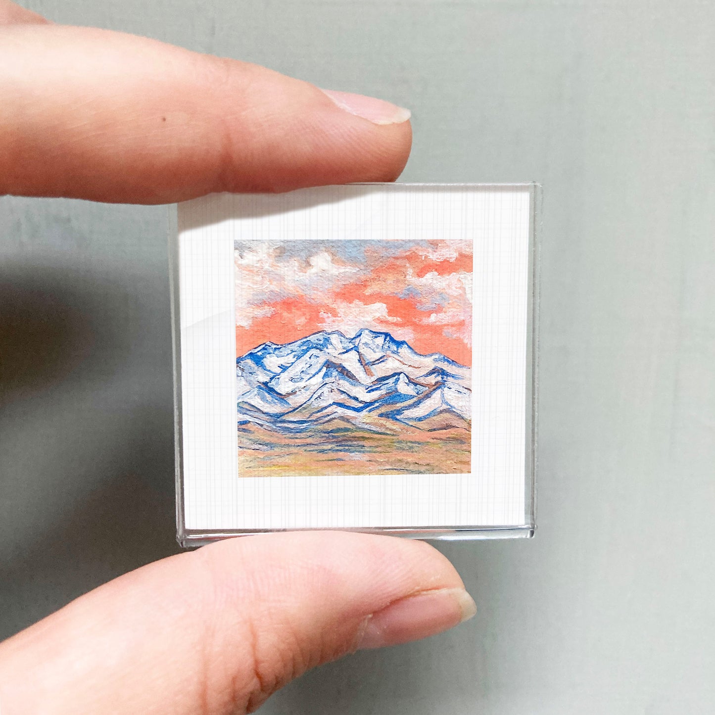 A person's hand is holding a small square picture of a mountain landscape with a colorful sky.