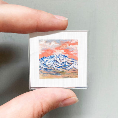A person's hand is holding a small square picture of a mountain landscape with a colorful sky.