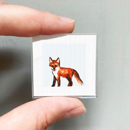 A person's hand is holding a small square with a picture of a fox on it.