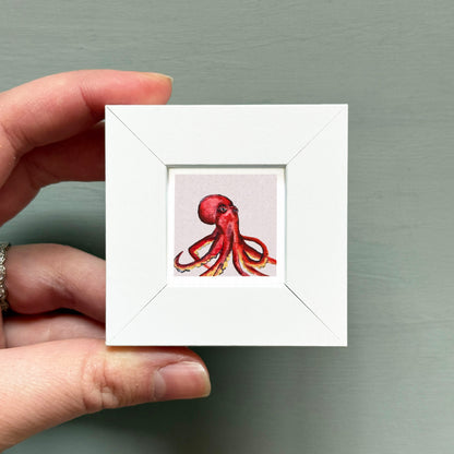 A hand is holding a small white frame containing a painting of an octopus.
