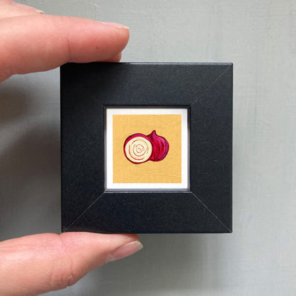 A person's hand is holding a small black square frame containing a framed illustration of a red onion.