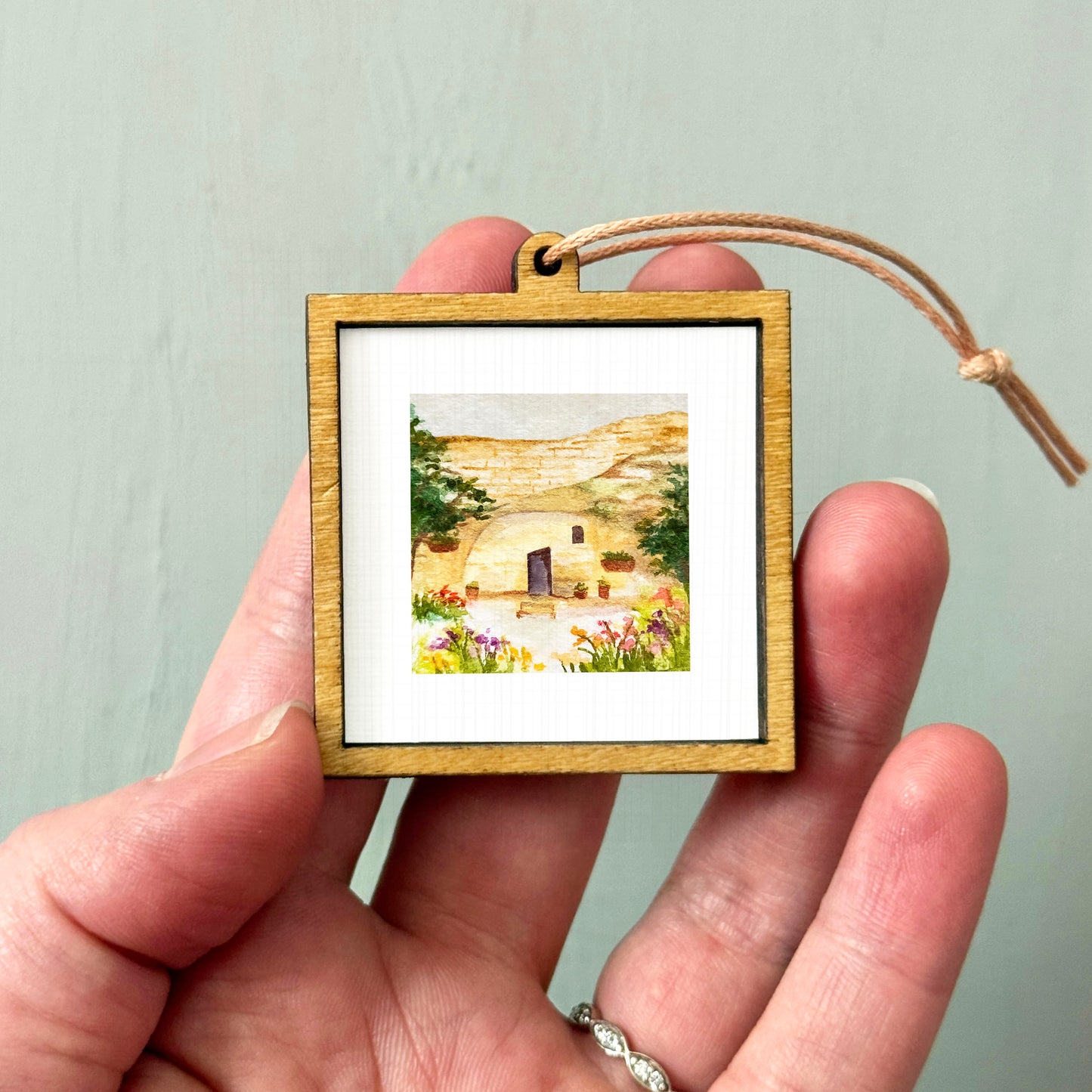 A hand holding a small wooden frame containing a watercolor painting of a desert landscape with a house and trees.