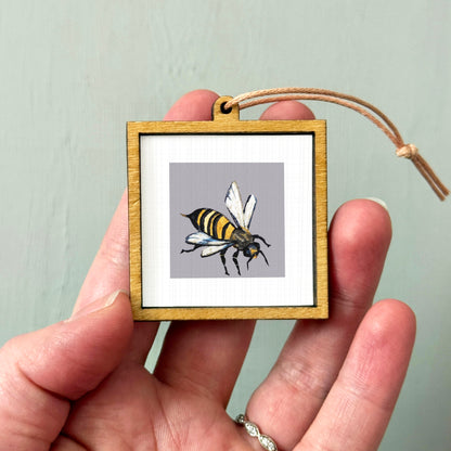 A person's hand holding a small wooden frame containing a picture of a bee.