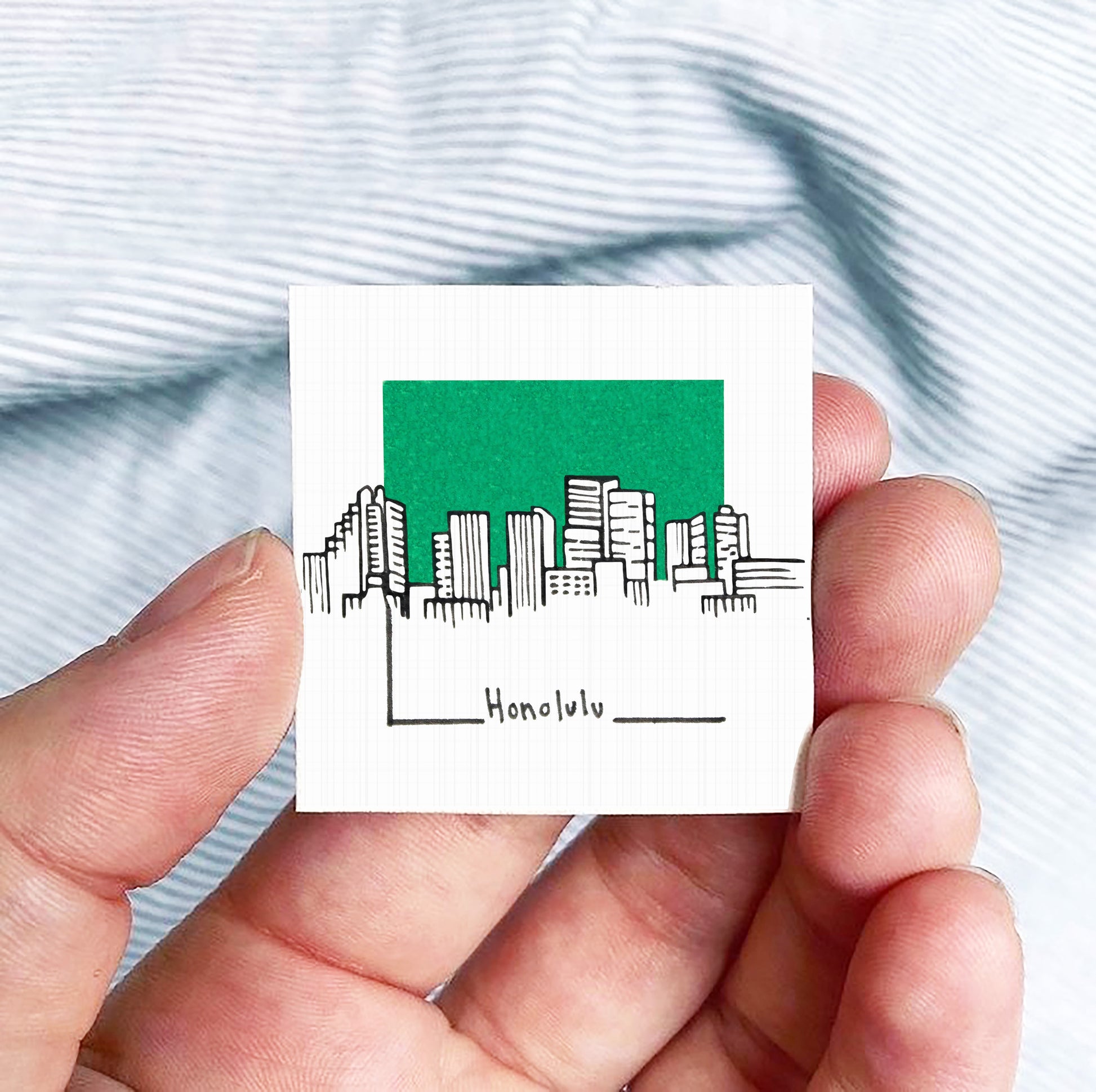 A hand holding a small square-shaped object with a green background depicting a city skyline and the word " Honolulu" written below it.