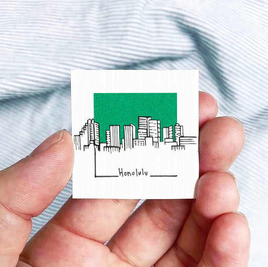 A hand holding a small square-shaped object with a green background depicting a city skyline and the word " Honolulu" written below it.