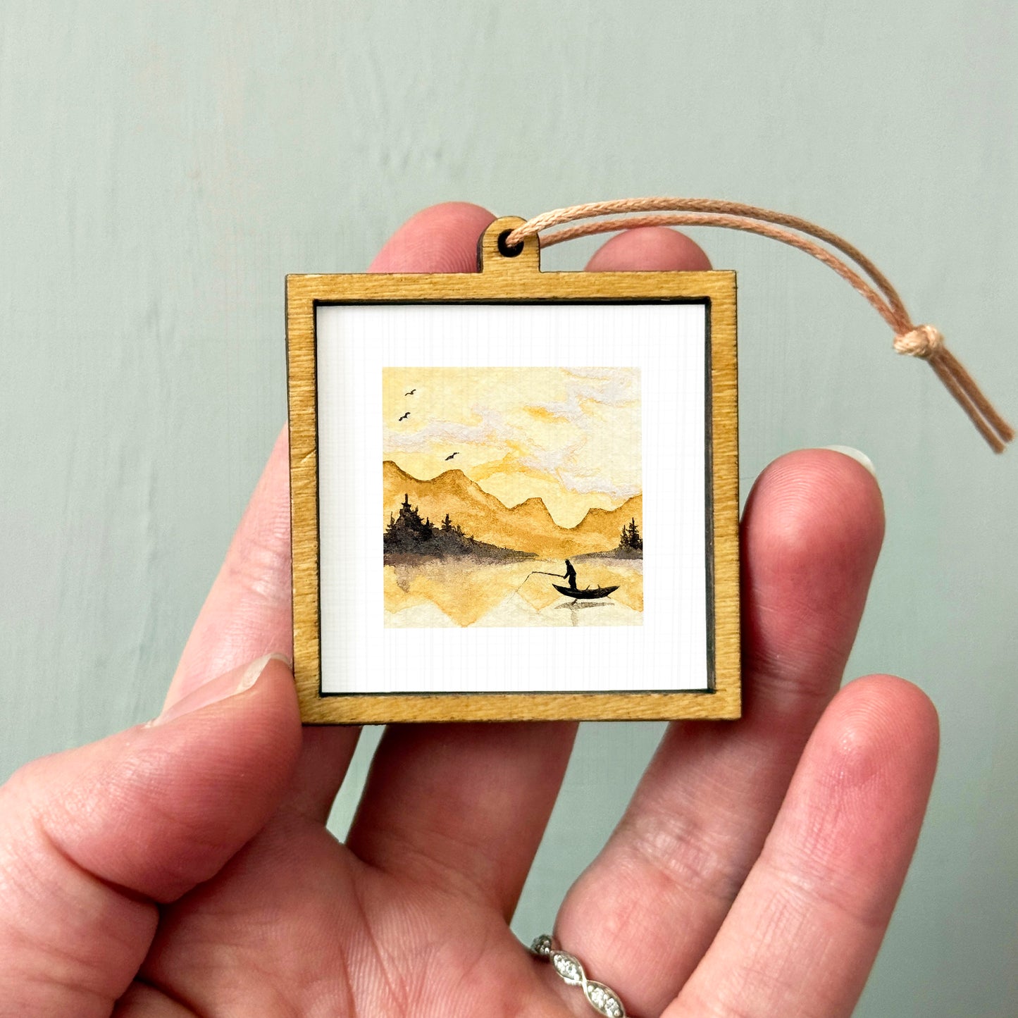 A hand holding a small wooden frame containing a watercolor painting of a landscape with mountains and a boat on a lake.