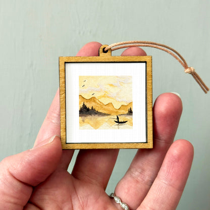 A hand holding a small wooden frame containing a watercolor painting of a landscape with mountains and a boat on a lake.