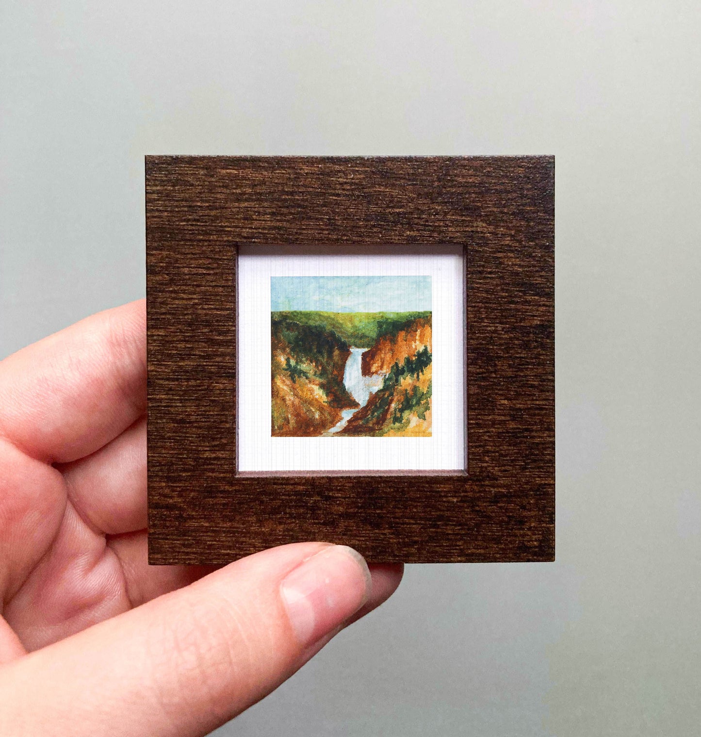 A hand is holding a small, square-shaped wooden frame containing a painting of a landscape with a river and mountains.