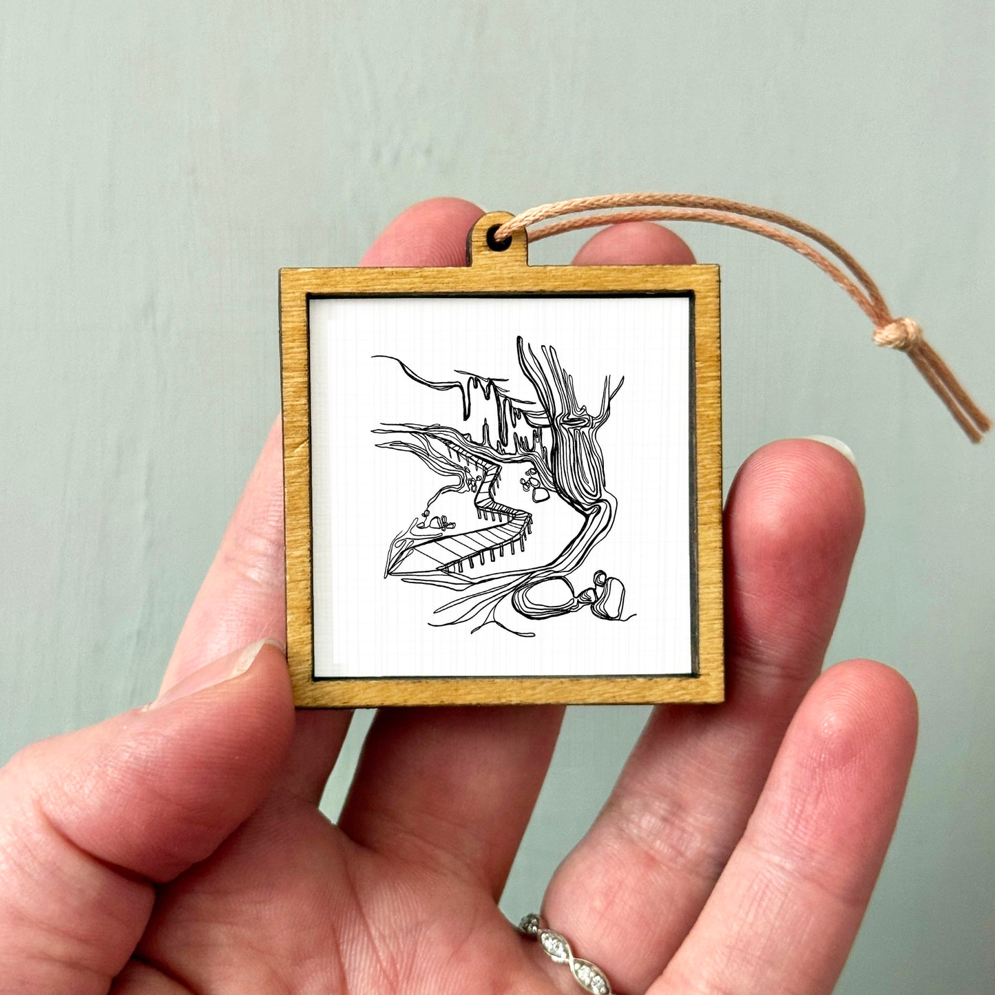 A hand holding a small wooden frame containing a black and white illustration of a bird or bird-like creature.