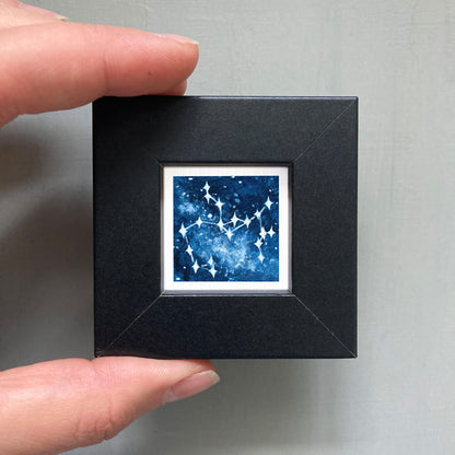 A hand is holding a small black square frame containing a framed image of a starry night sky.