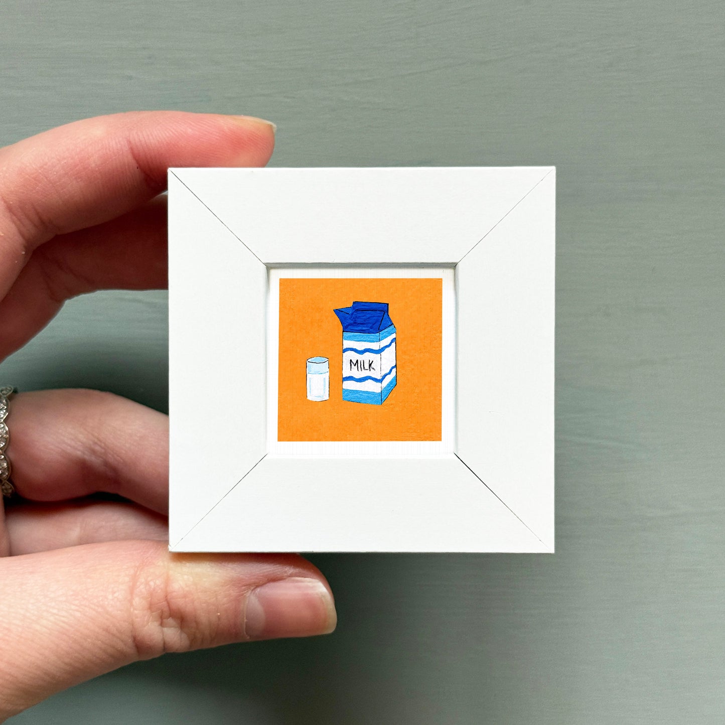 A hand is holding a small white picture frame containing an illustration of a carton of milk and a glass.