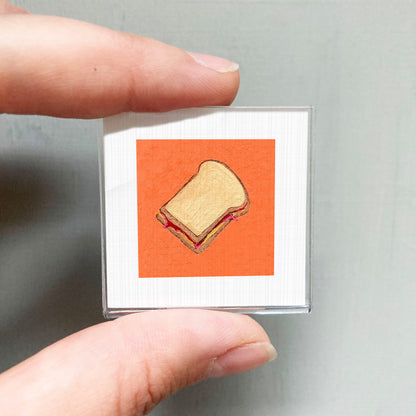 A person's hand holding a small square with an illustration of a sandwich on it.