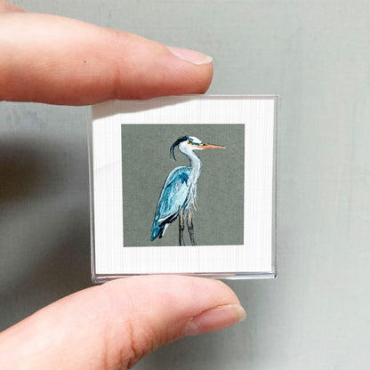 A person's hand is holding a small square picture of a blue heron.
