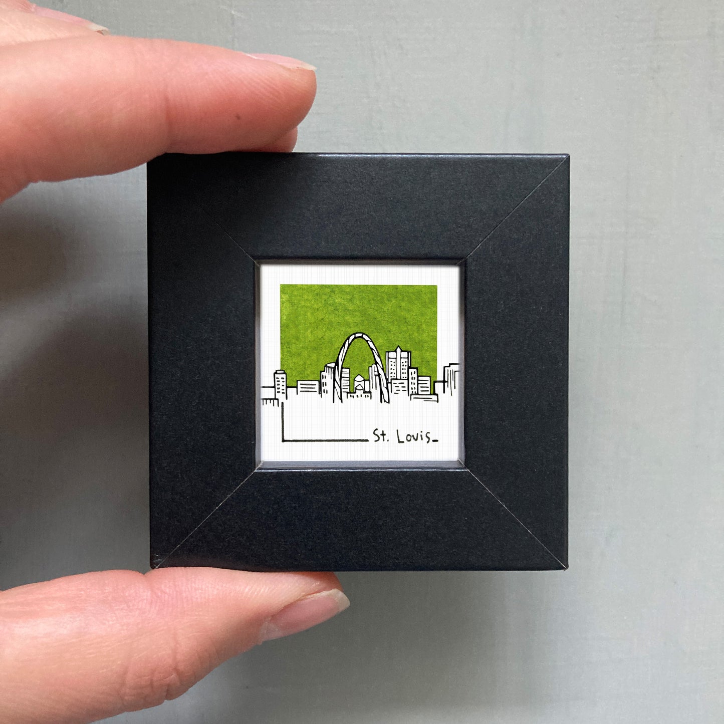 A person's hand holding a small black square frame containing a miniature illustration of St. Louis, Missouri.