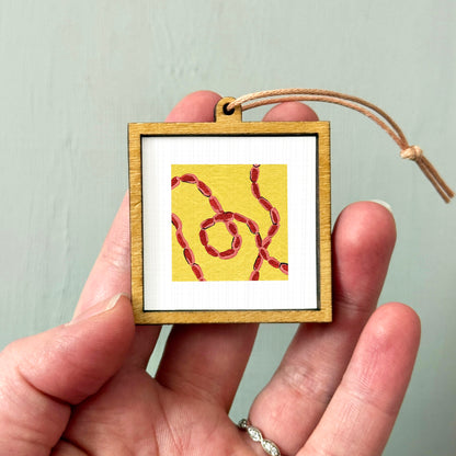 A hand holding a small wooden frame containing a framed artwork with a yellow background and red lines.
