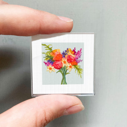 A hand is holding a small square card with a vibrant watercolor painting of a colorful bouquet of flowers.
