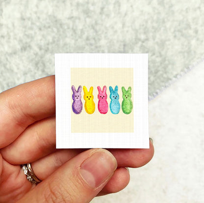 A hand holding a small square card with a colorful illustration of five Easter bunny characters on it.