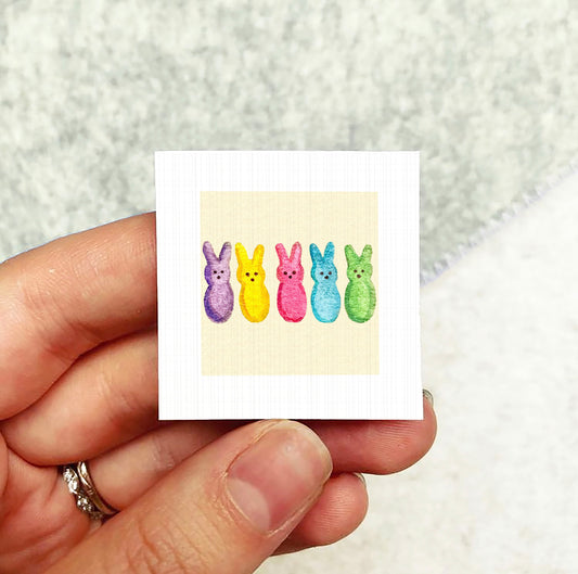 A hand holding a small square card with a colorful illustration of five Easter bunny characters on it.