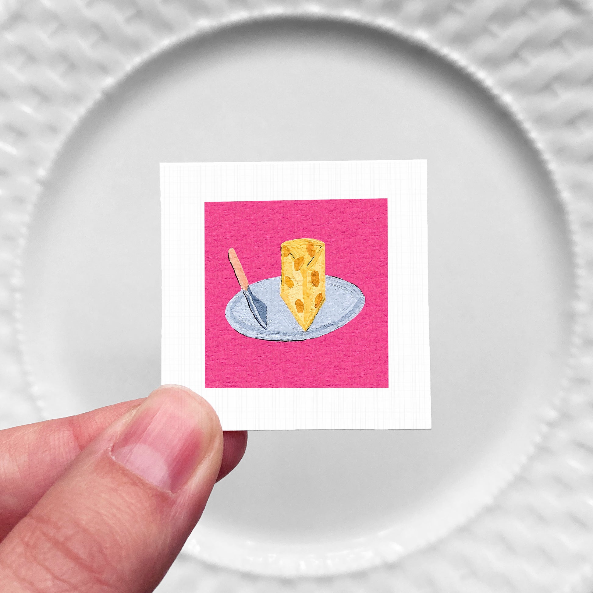 A hand is holding a small square with a pink background and a yellow cheese slice on a white plate.