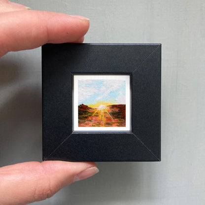 A person's hand holding a small, square-shaped black frame containing a miniature painting of a landscape with a sunset.