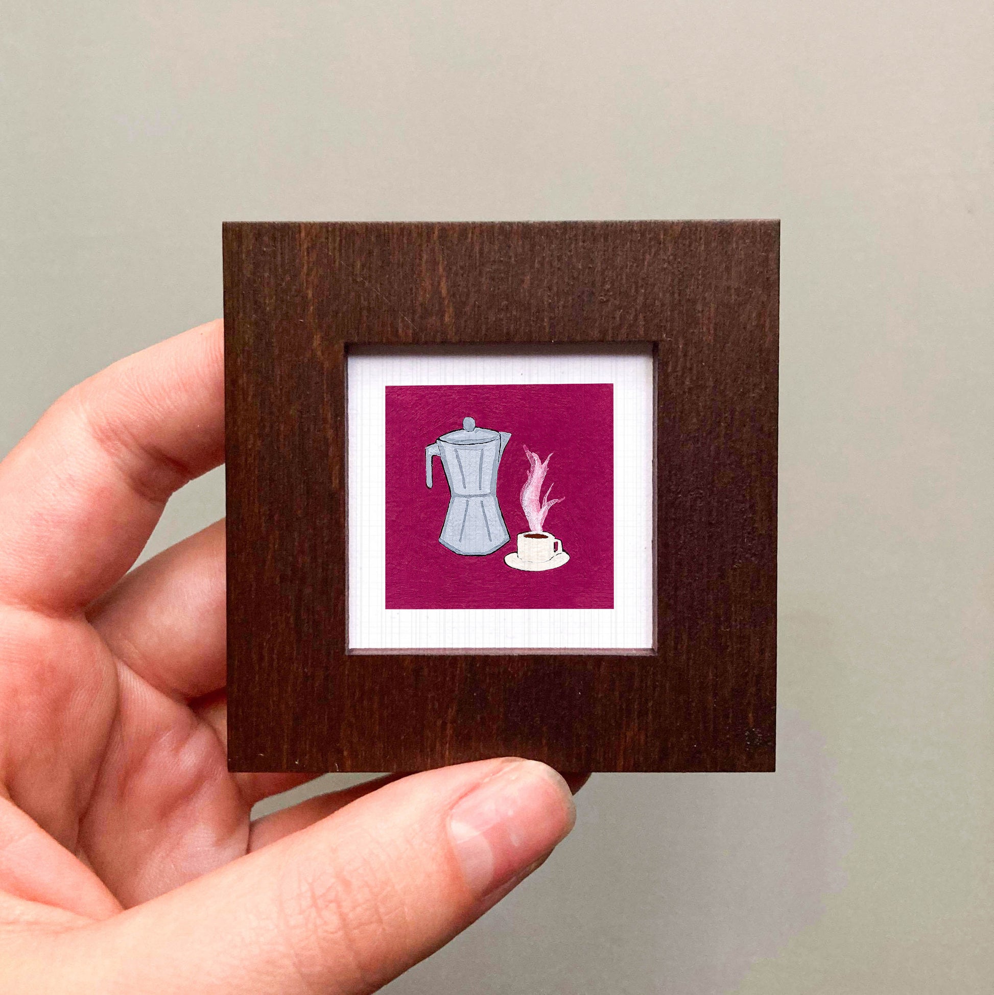 A hand is holding a small square frame containing an illustration of a coffee maker and a cup of coffee.