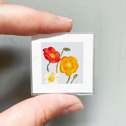 A person's hand holding a small square picture of three colorful flowers, including red, yellow, and white flowers.