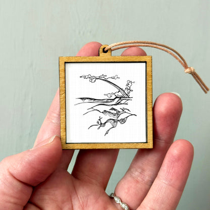 A hand holding a small wooden frame containing a black and white illustration of a landscape with mountains and clouds.