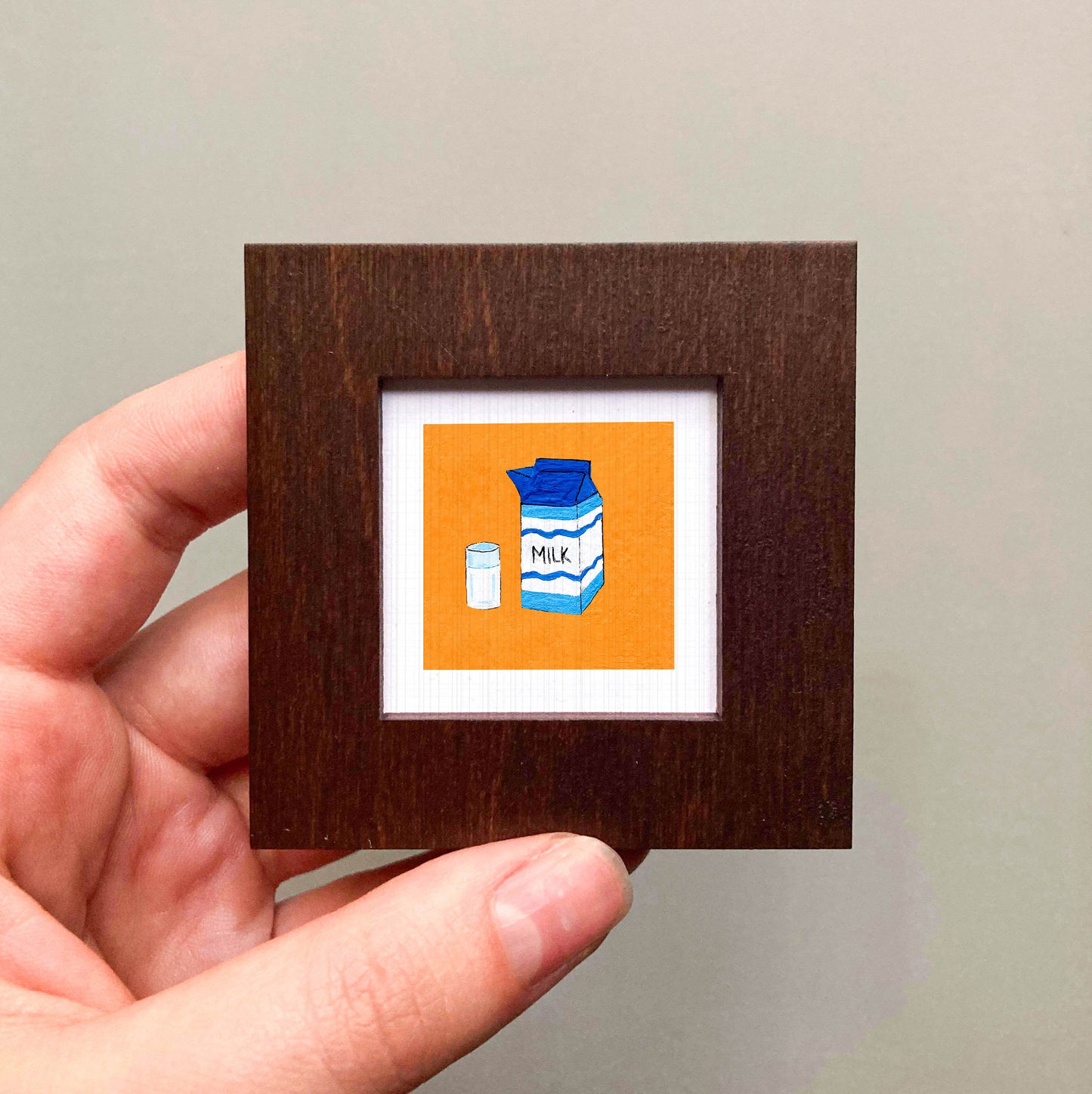 A hand is holding a small square frame containing an illustration of a carton of milk and a glass of milk.