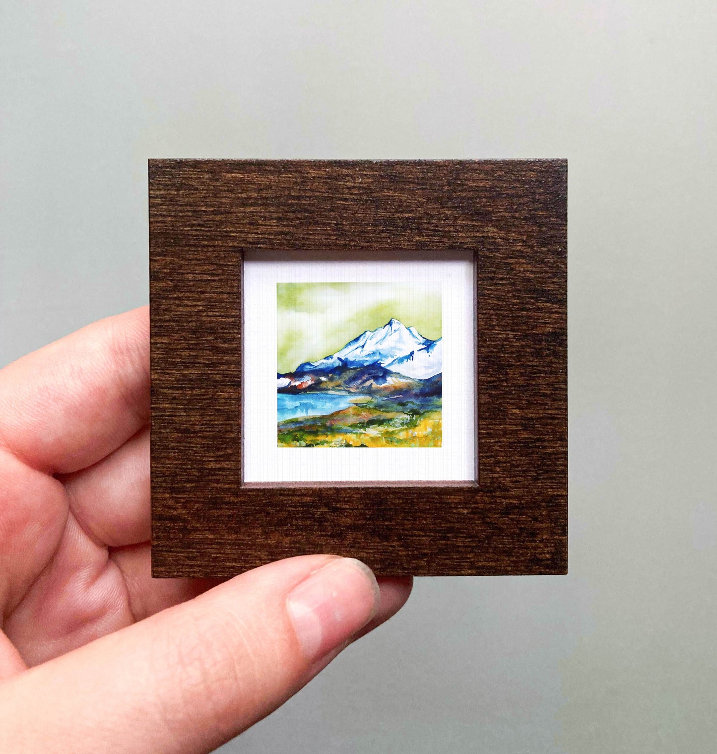 A hand is holding a small, square-shaped wooden frame containing a watercolor painting of a mountain landscape.