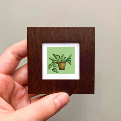 A hand is holding a small wooden frame containing a framed painting of a potted plant with green leaves.