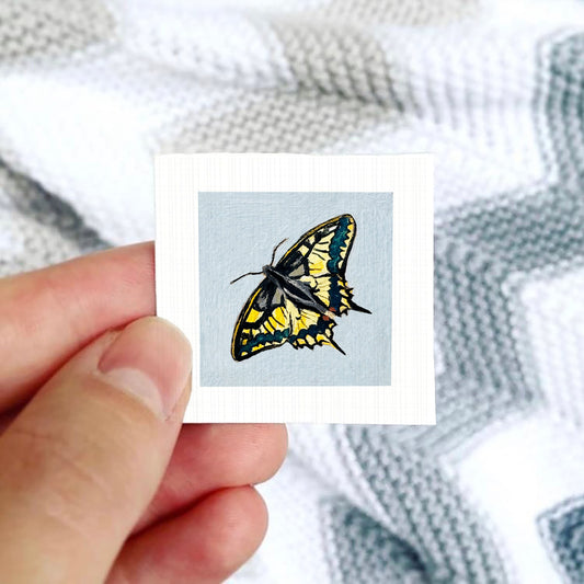 A hand is holding a small square card with a colorful butterfly illustration on it.