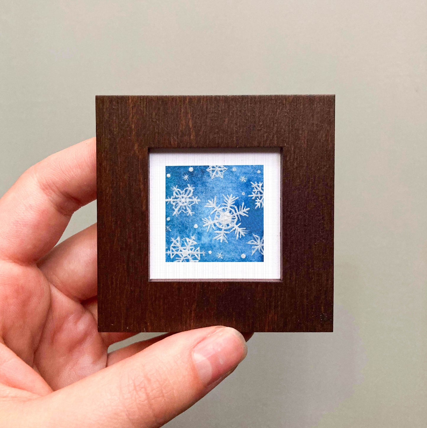 A hand is holding a small square picture frame containing a blue and white snowflake design.