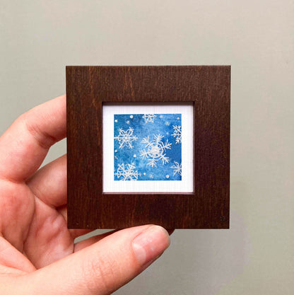 A hand is holding a small square picture frame containing a blue and white snowflake design.