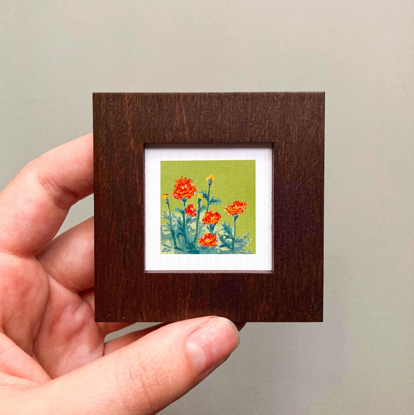 A hand is holding a small wooden frame containing a framed painting of red flowers against a green background.