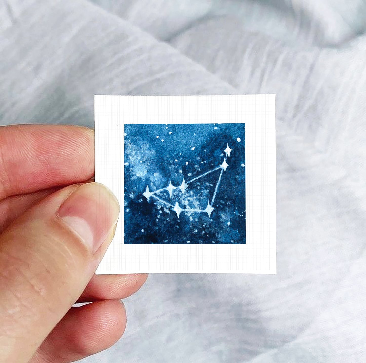 A hand is holding a small square-shaped paper with a depiction of a starry night sky on it.