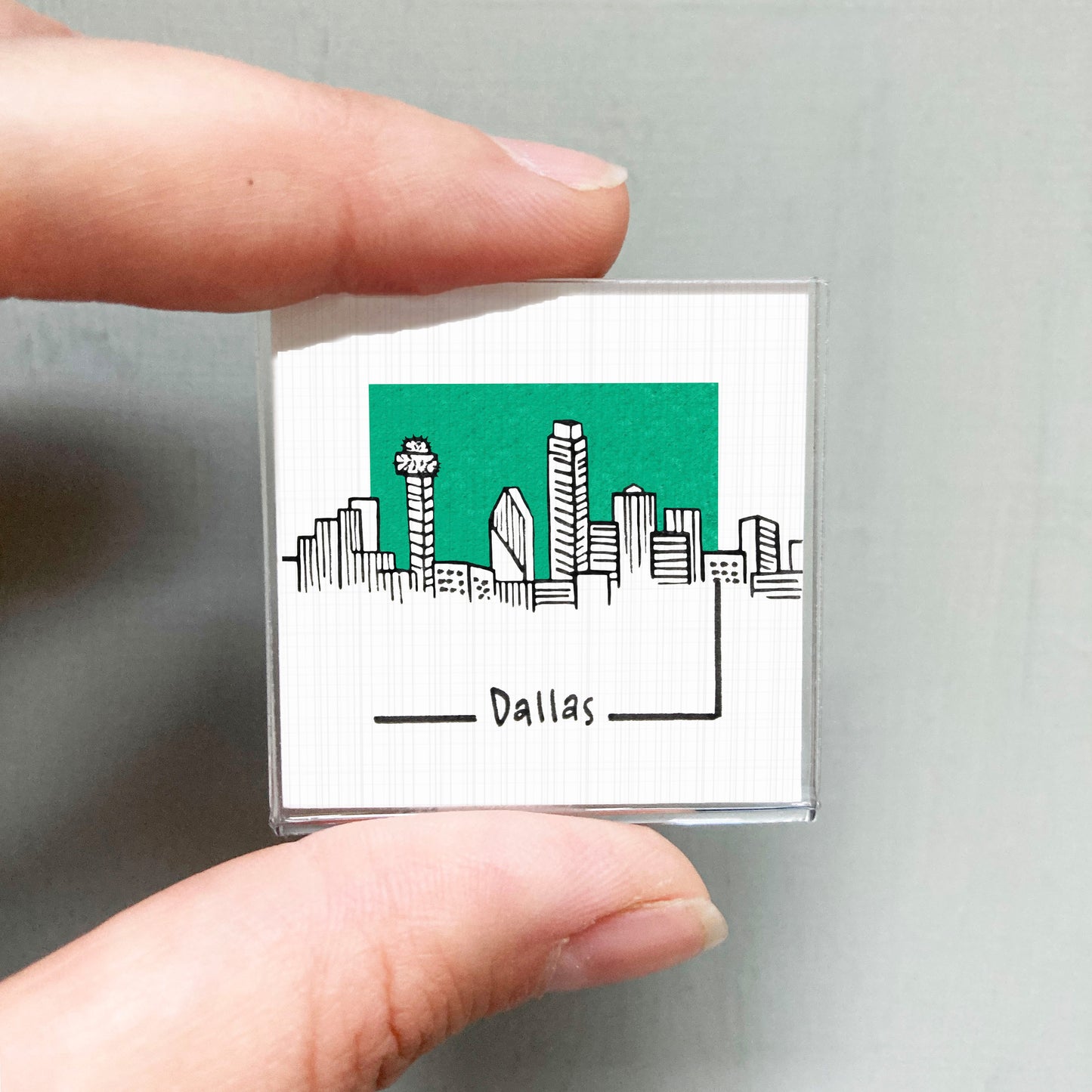 A hand holding a small square with a drawing of a city skyline and the word "Dallas" written below it.
