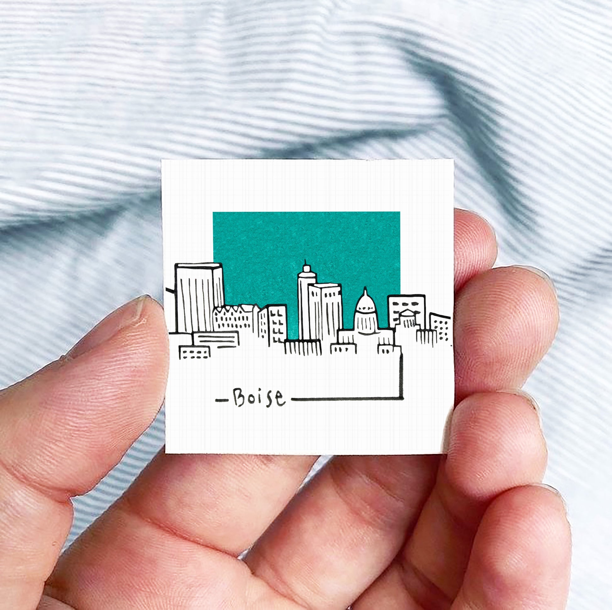 A hand holding a small square-shaped object with a drawing of a city skyline on it, against a striped background.