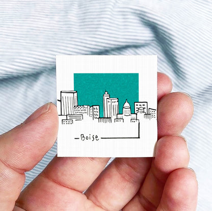 A hand holding a small square-shaped object with a drawing of a city skyline on it, against a striped background.