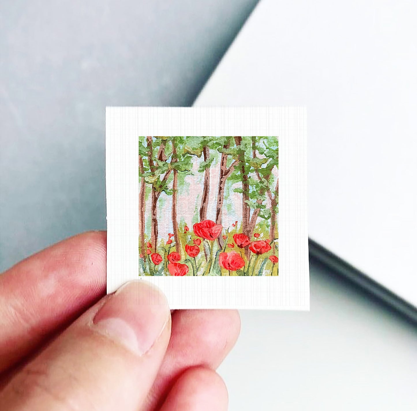 A hand holding a small square card with a painting of red flowers and trees on it.