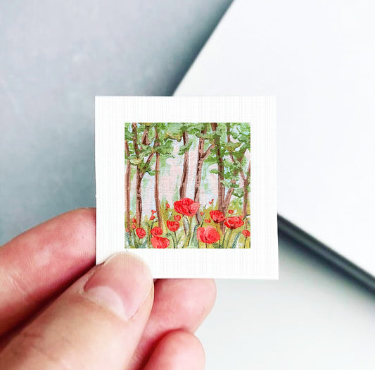 A hand holding a small square card with a painting of red flowers and trees on it.