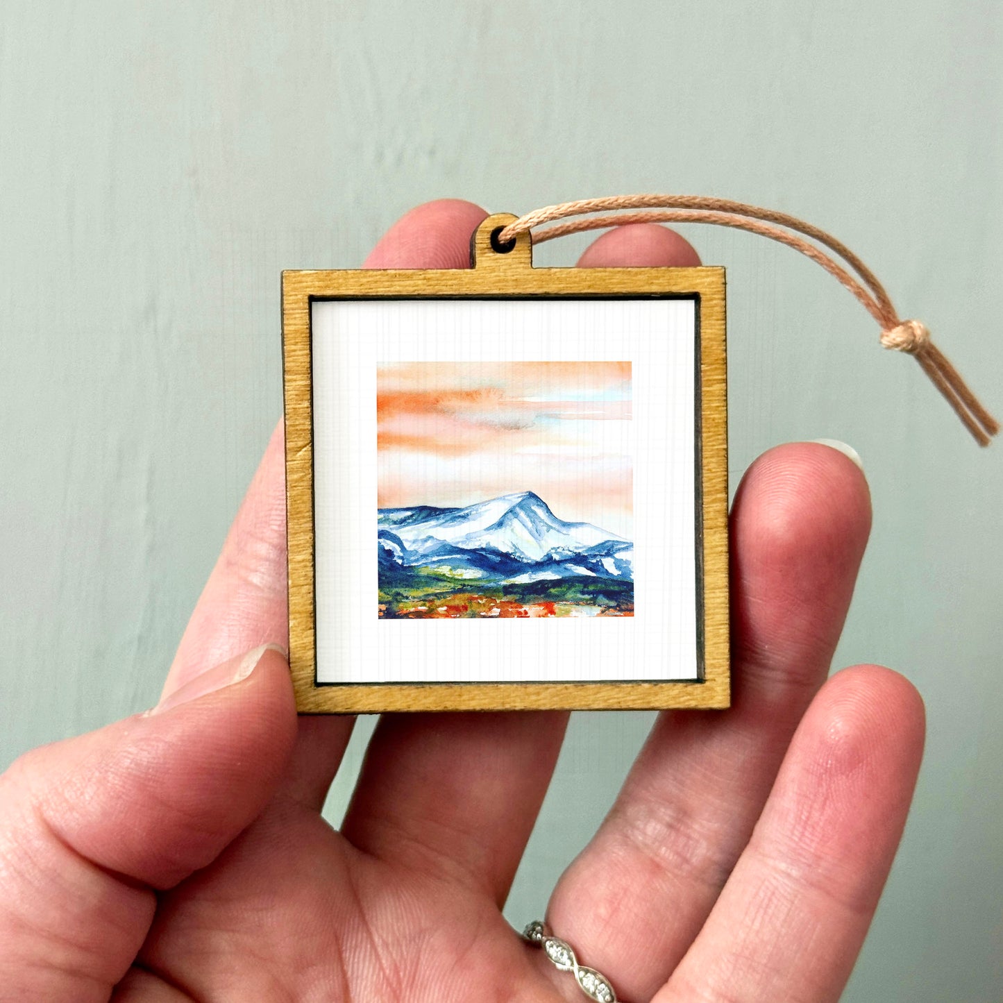 A hand holding a small wooden frame containing a watercolor painting of a mountain landscape.