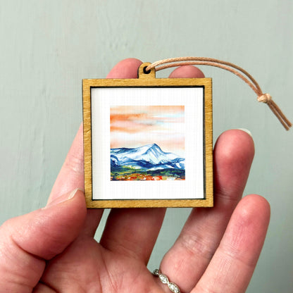 A hand holding a small wooden frame containing a watercolor painting of a mountain landscape.