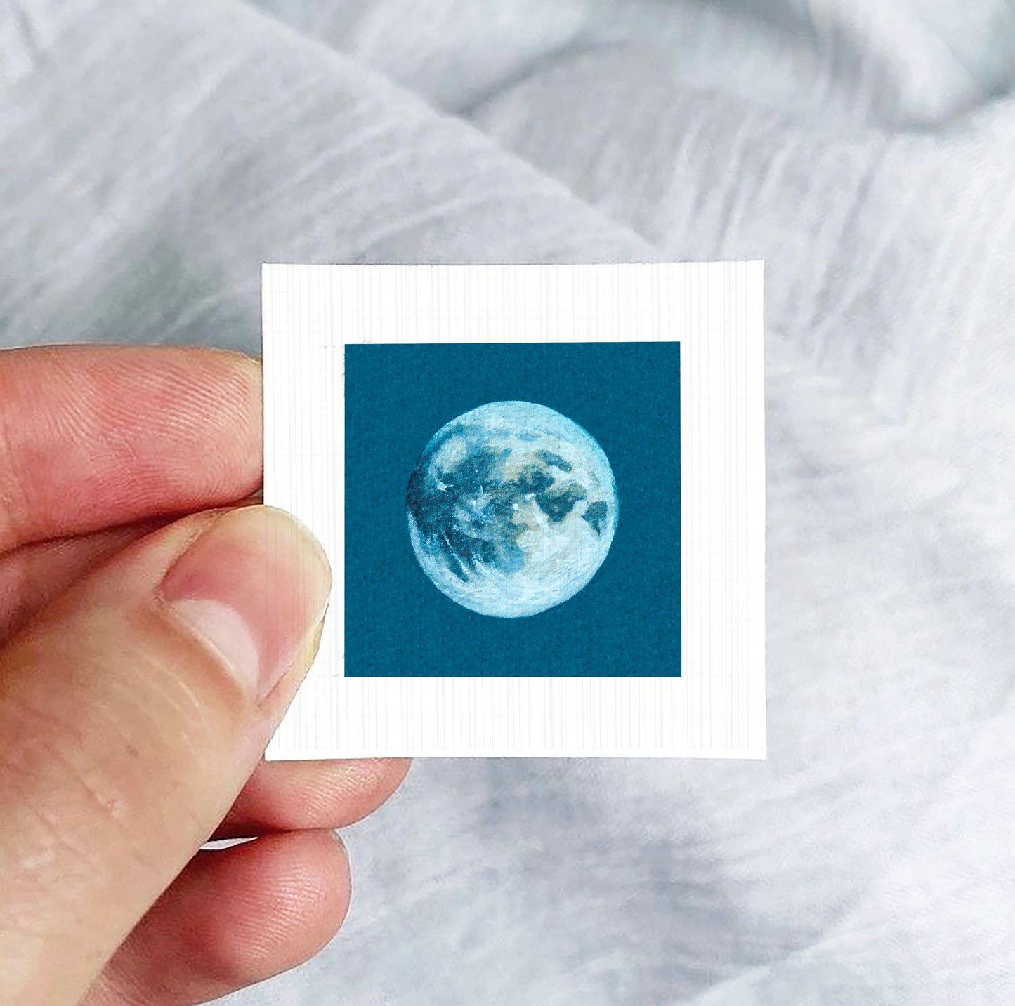 A hand is holding a small square-shaped paper with a depiction of the moon on it.
