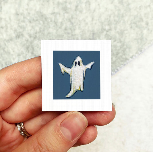 A hand holding a small square picture of a ghost against a blue background.