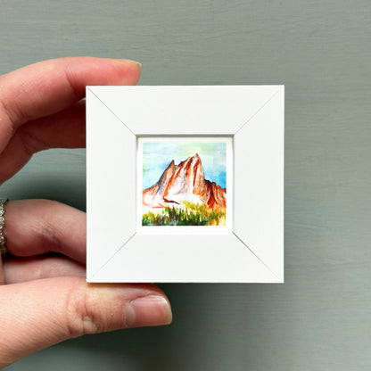 A hand is holding a small white square frame containing a watercolor painting of a mountain landscape.