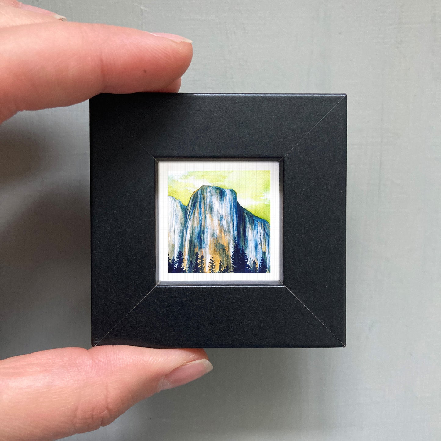 A person's hand holding a small black frame containing a framed painting of a mountain landscape.