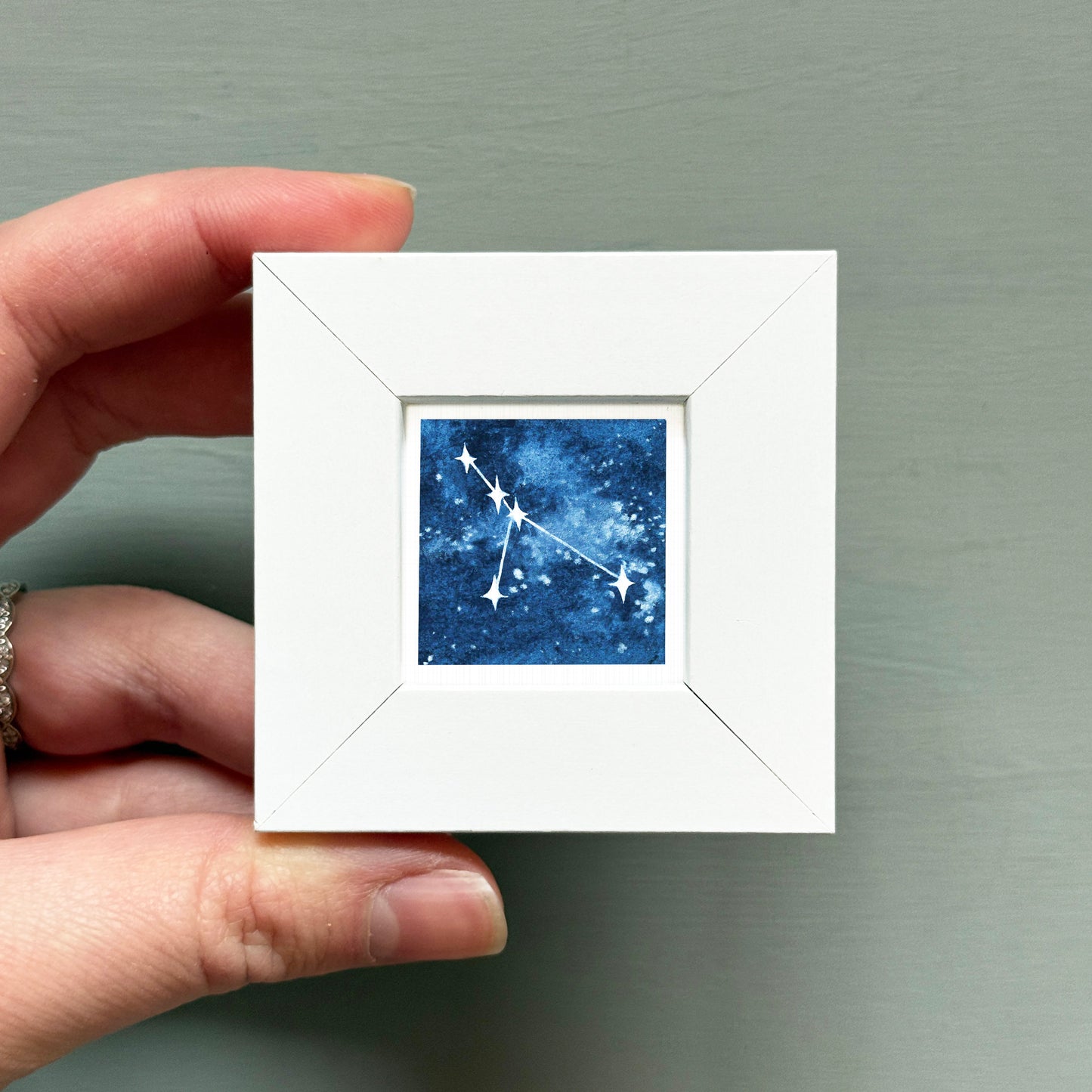 A hand is holding a small white square frame containing a depiction of the constellation of Cancer.