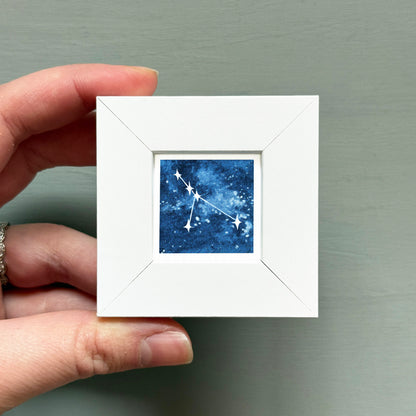 A hand is holding a small white square frame containing a depiction of the constellation of Cancer.