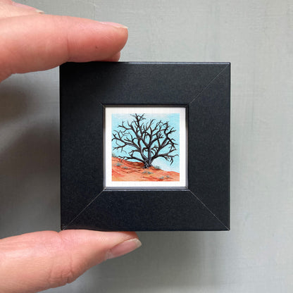 A person's hand is holding a small black frame containing a framed painting of a tree with bare branches against a blue sky.